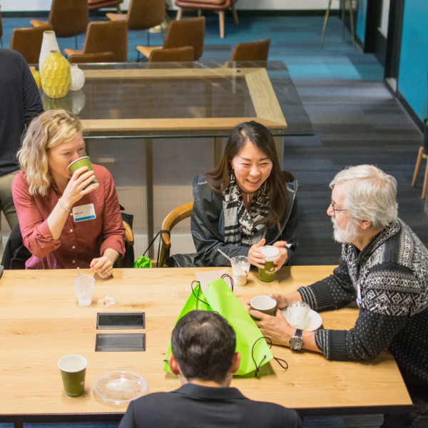 Two men and two women seated around a wooden table, drinking from paper cups and laughing together