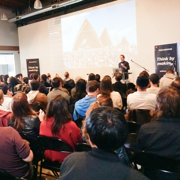 Man with a presentation screen behind him presenting and speaking to a large, seated crowd